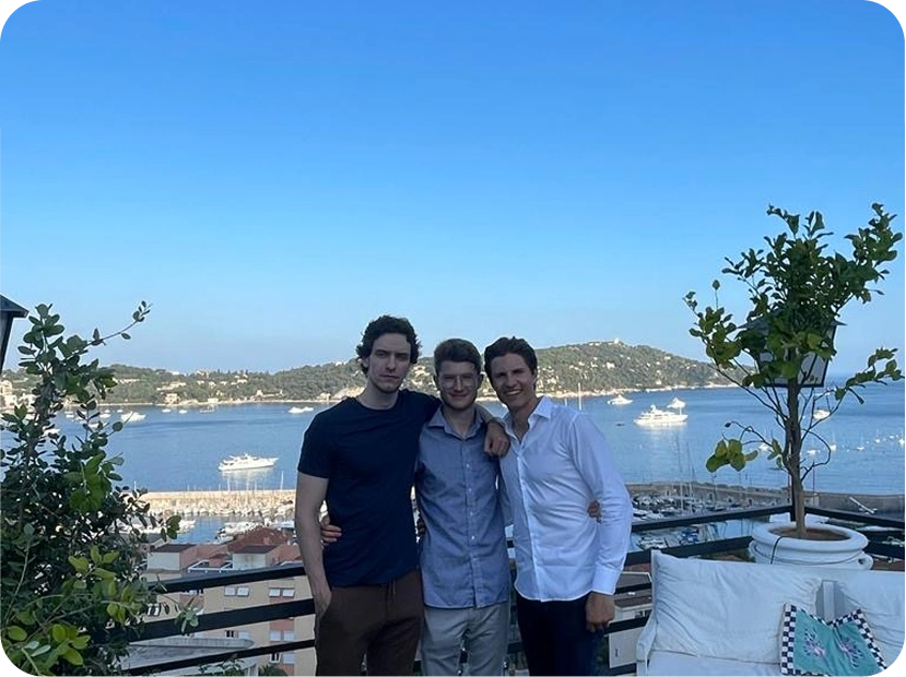 Three men posing together on a balcony overlooking a marina with boats and hills in the background.