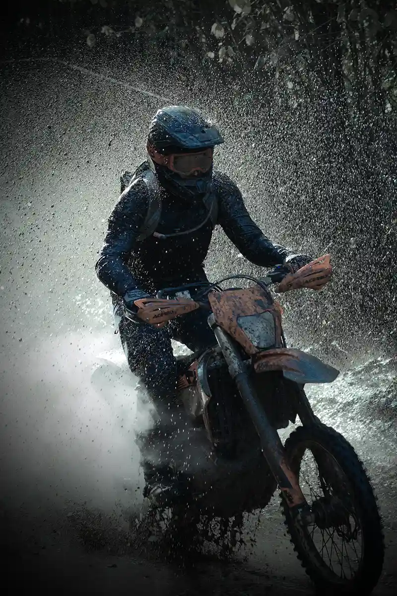 Motorcyclist in helmet riding dirt bike through water, spraying droplets in wooded area.