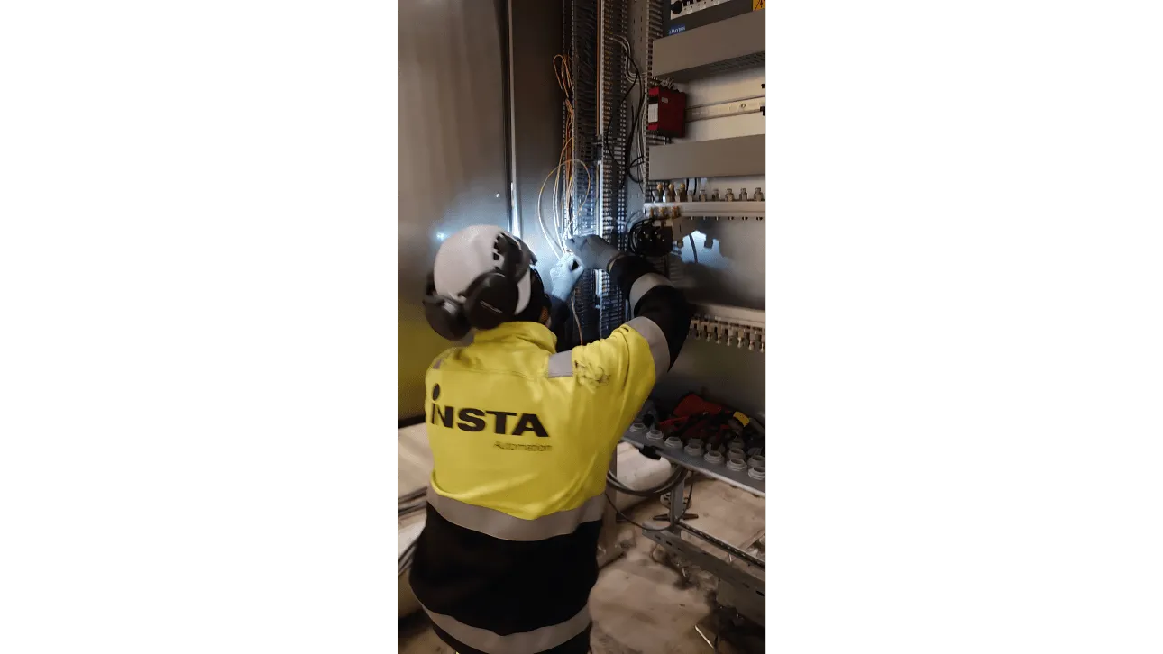 Henkilö työskentelee sähkökaapin sisällä kytkien johtoja. Hänellä on yllään keltainen ja musta huomioväritakki, valkoinen suojakypärä, kuulosuojaimet ja käsineet. Työskentelyaluetta valaisee kirkas valo, joka heijastuu johdoista.