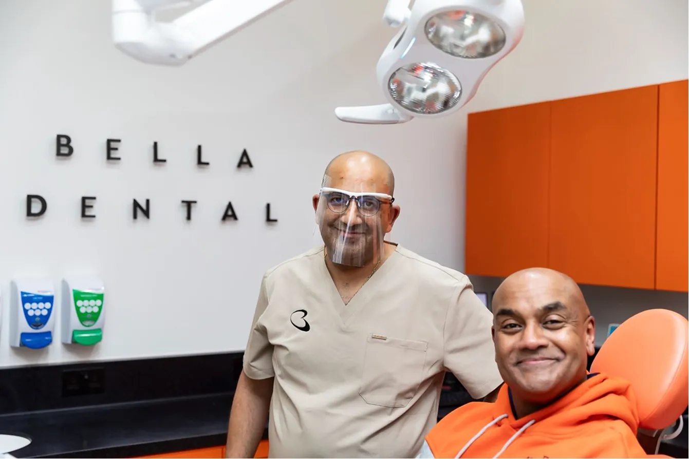 Dentist standing beside a smiling patient seated in the dental chair, with the Bella Dental sign behind them