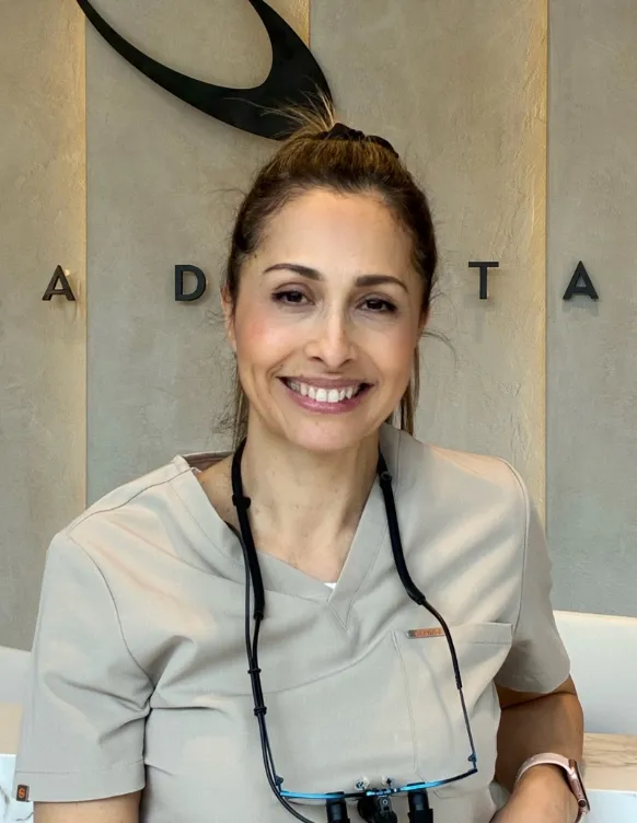 Smiling woman with light brown hair tied back wearing a beige medical scrub top and magnifying glasses around her neck, sitting in front of a wall with partial letters visible.