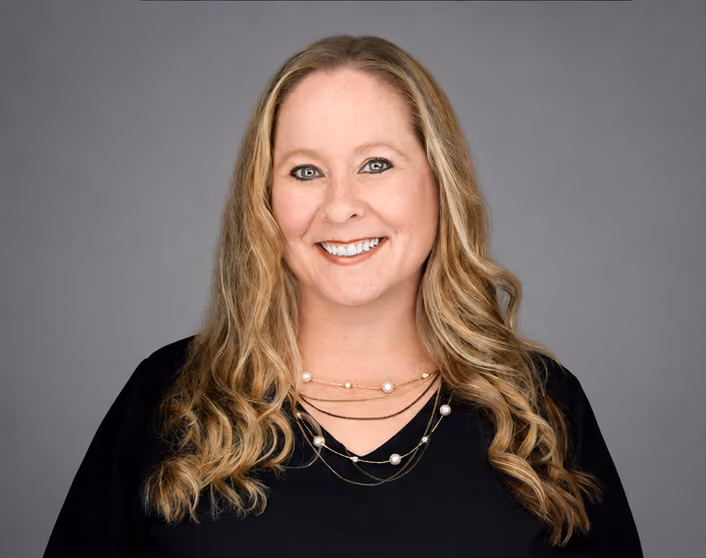 A headshot of Becky DuLac with long blonde curly hair wearing a black top and layered pearl necklace against a gray background.