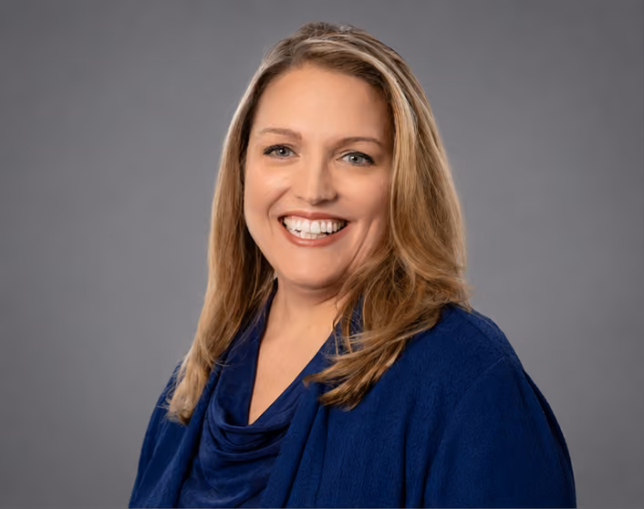 A headshot of Kristi Castro with blonde hair wearing a blue top and jacket against a plain gray background.