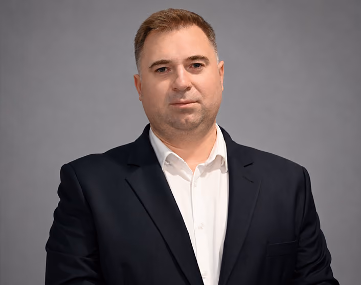 A headshot of Juliusz Czekaj in a white shirt and dark blazer posing against a gray background.