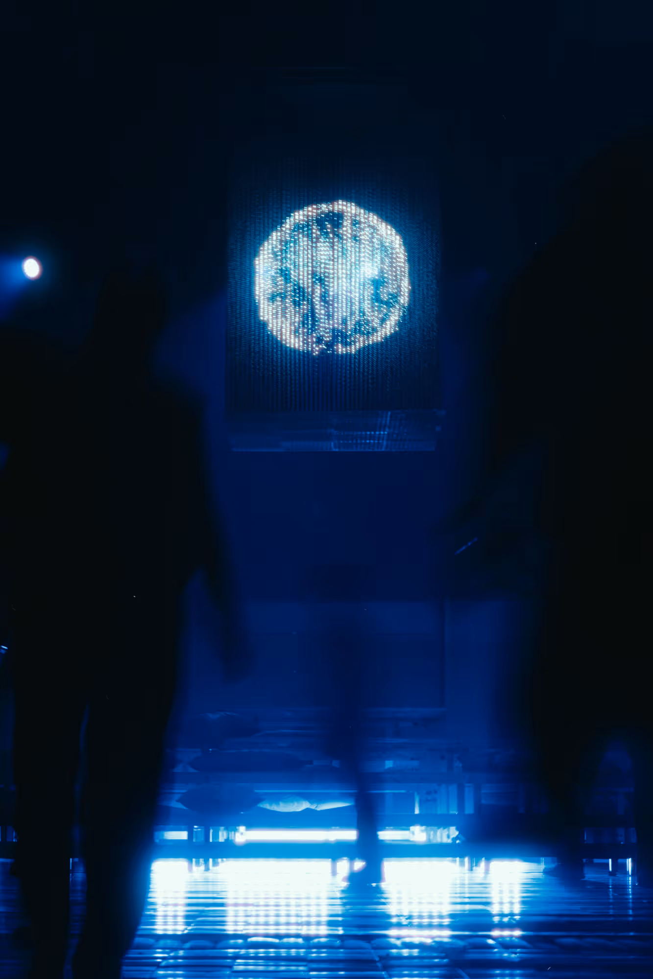 Blurred silhouettes of people walking in a dark room illuminated by a glowing circular display above them.