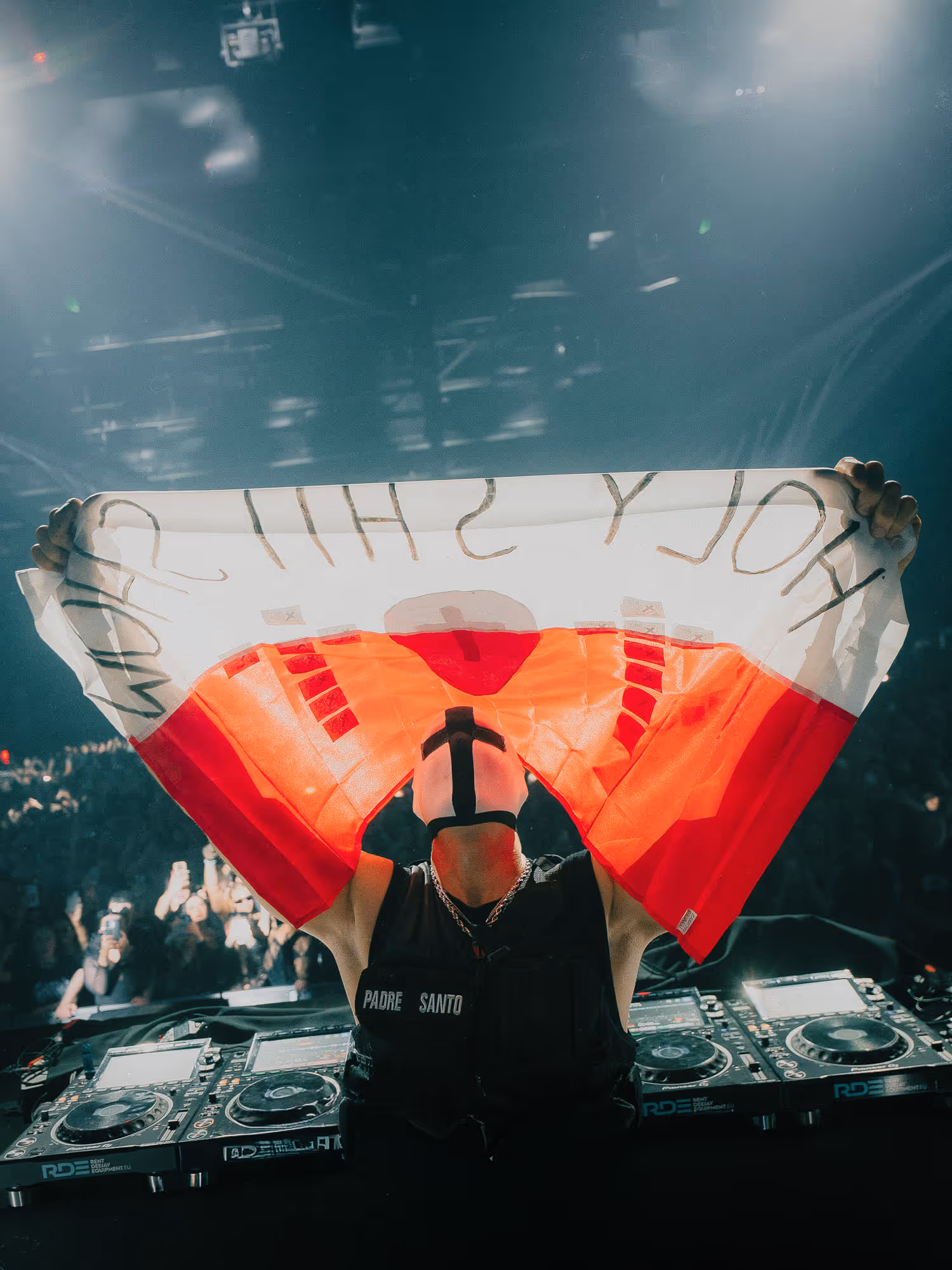 DJ wearing a mask with a cross holds a red and white flag reading Holy Shit at a concert.