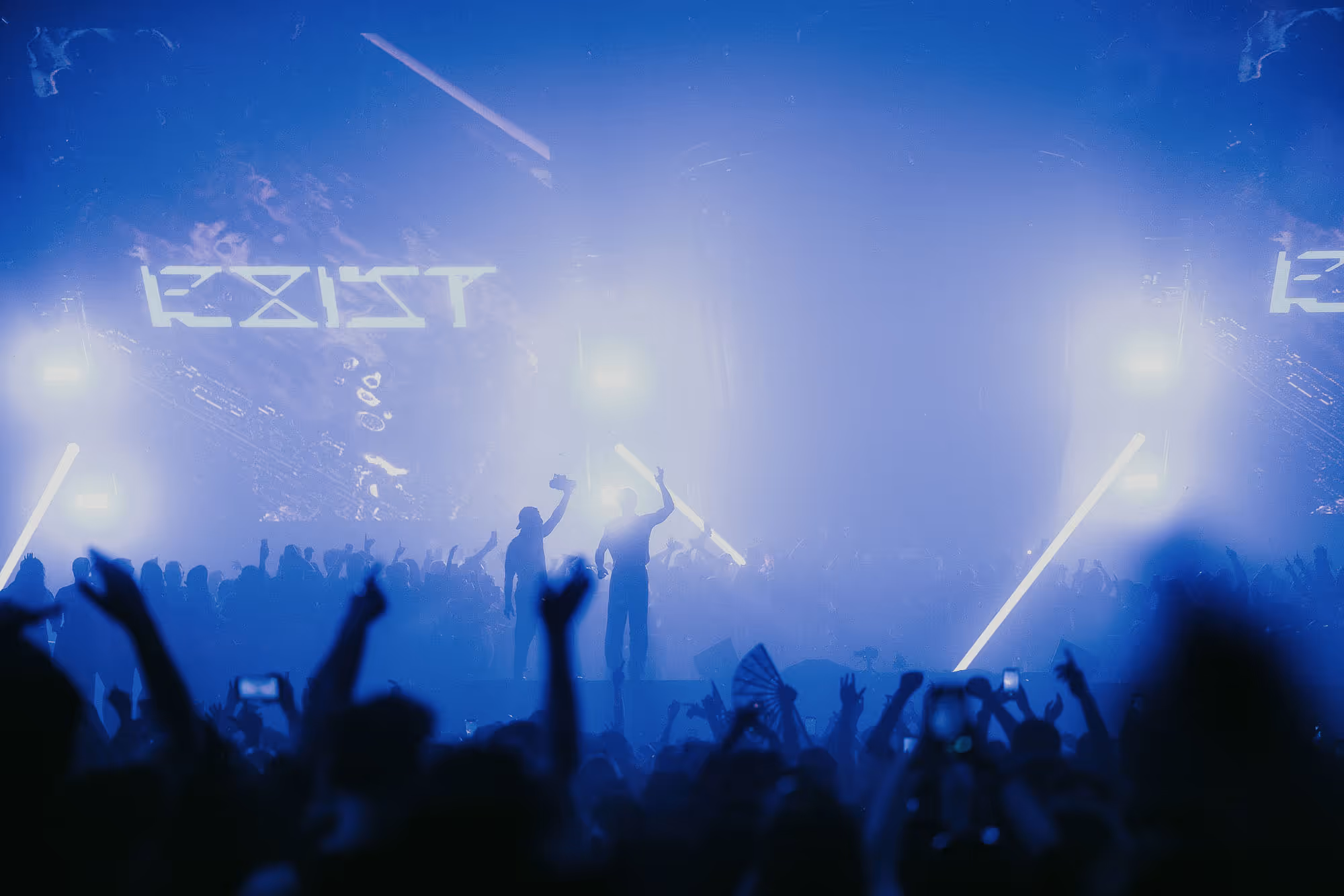 Crowd at a concert with two people on stage illuminated by blue lights and lasers.