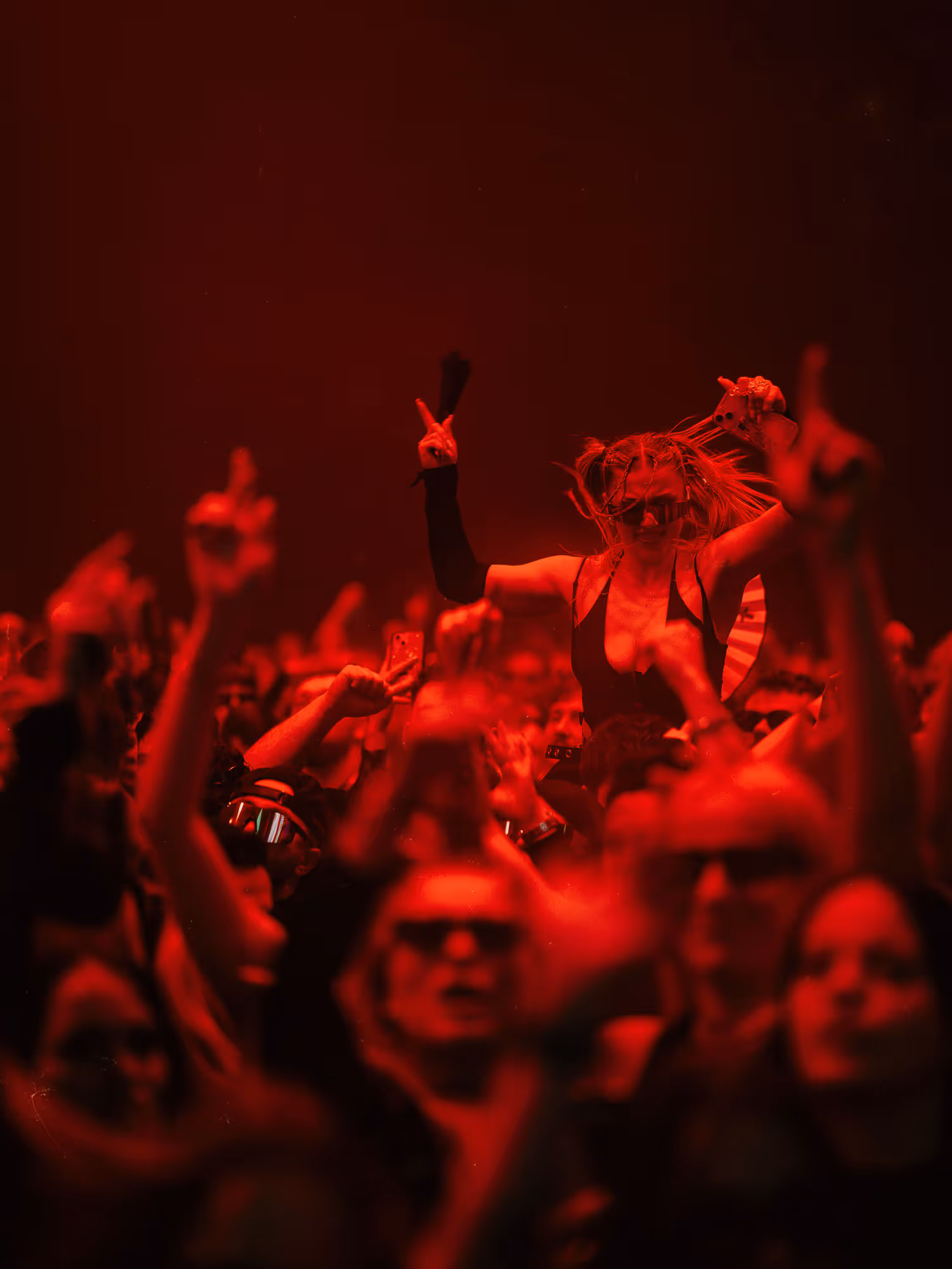 Crowd at a concert with a woman in sunglasses and braided hair raised above the crowd, red lighting.