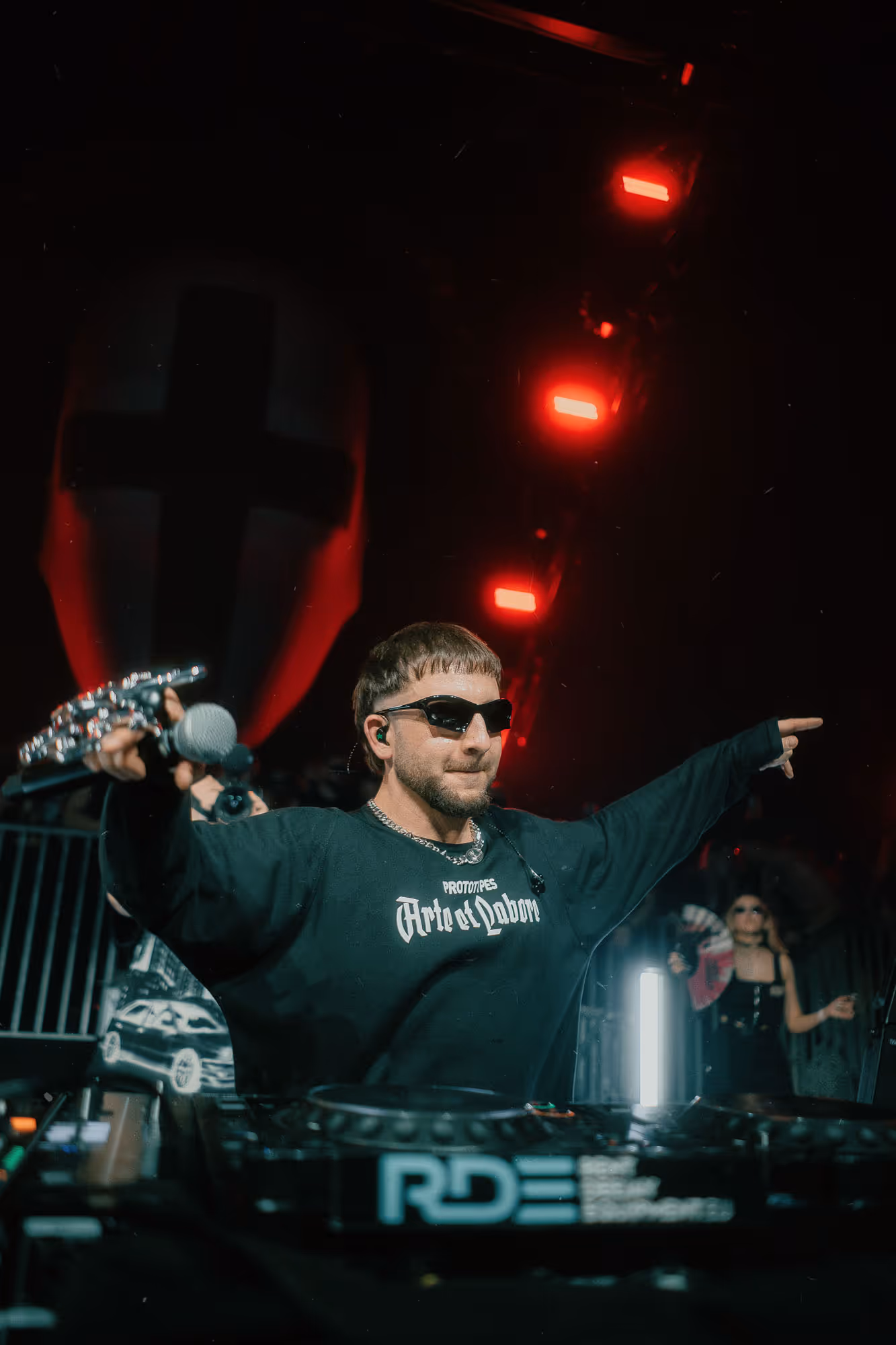 DJ wearing sunglasses and black shirt cheering with arms outstretched in front of mixing equipment and red stage lights.
