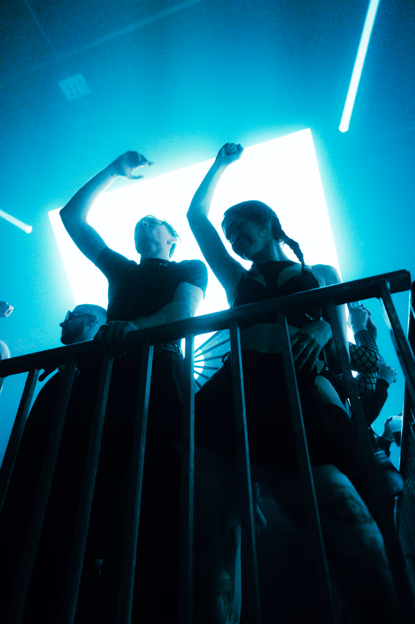 People dancing and raising arms near metal railing under bright blue light at a nightclub.