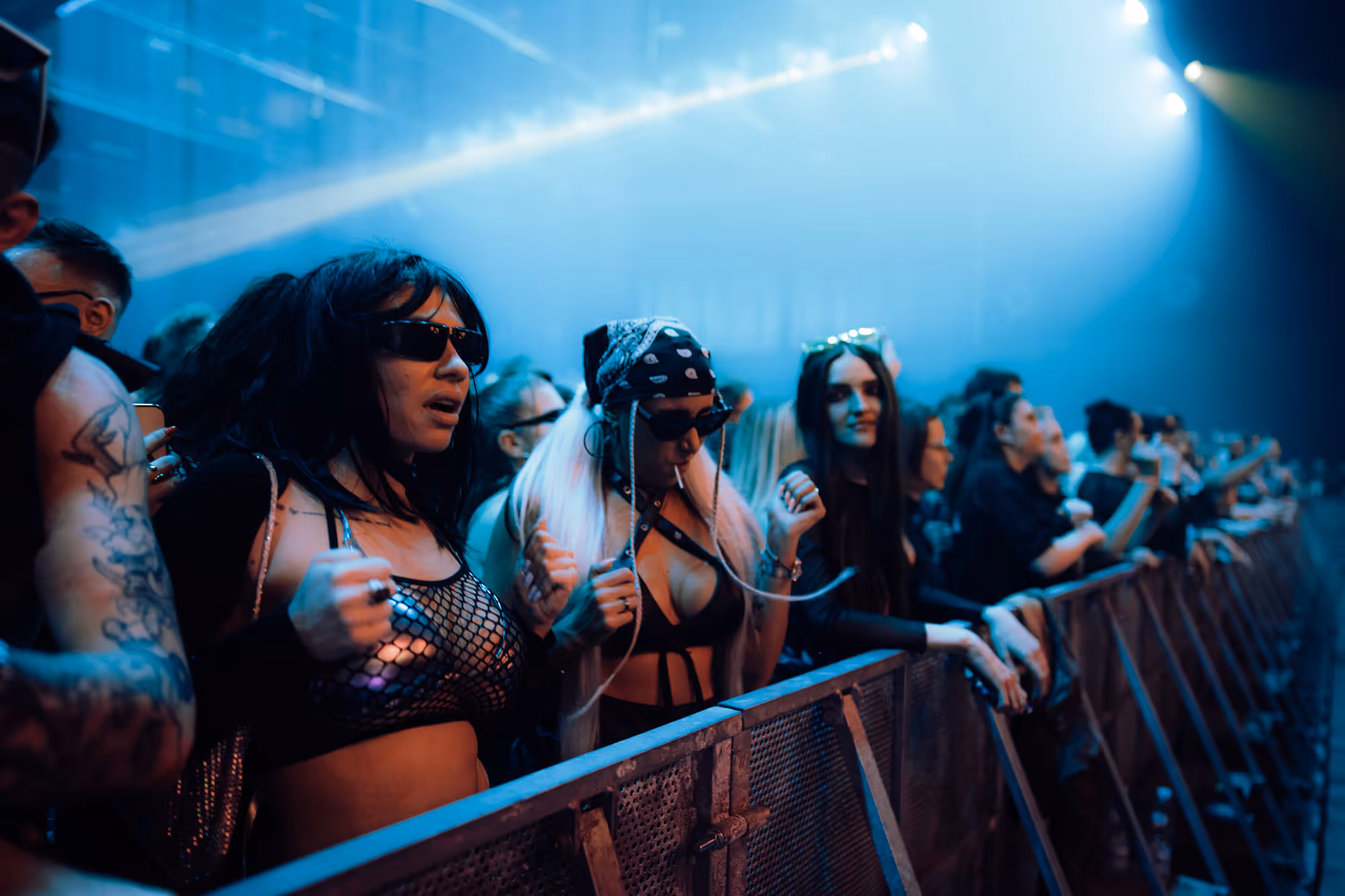 People in eclectic outfits enjoying a concert near a metal barrier under blue lighting.