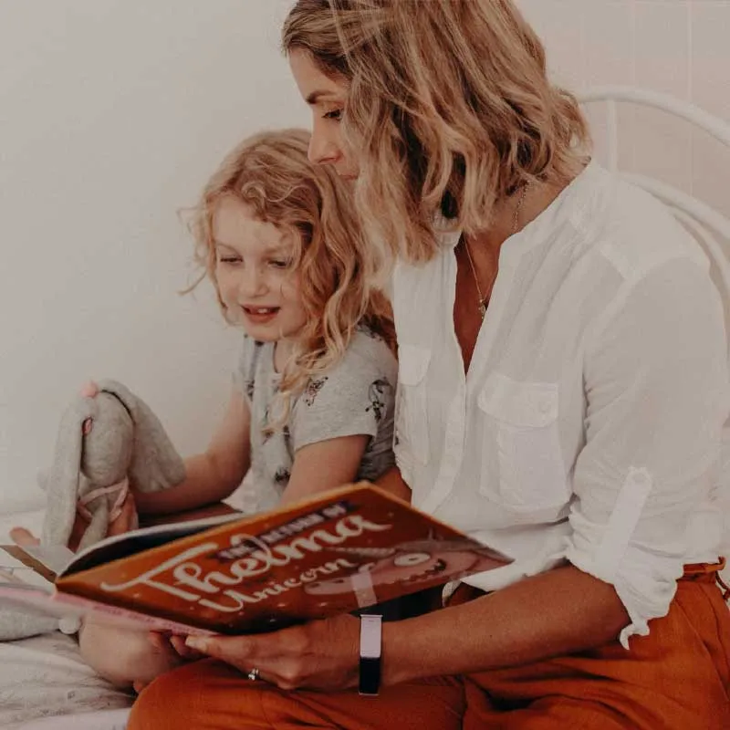 Woman reading a book titled Phelma Unicorn to a young girl holding a stuffed bunny.