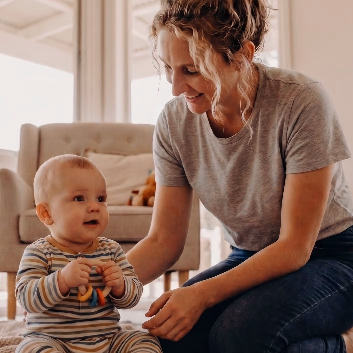 Smiling woman interacts with a seated baby holding a colorful ring toy in a cozy living room.