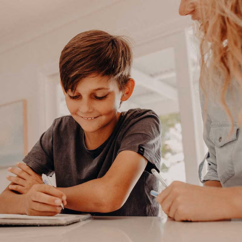Young boy smiling while writing with a woman assisting him at a table indoors.