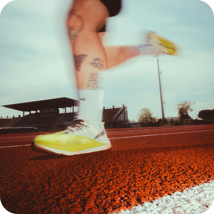 Legs of a runner with tattoos wearing yellow running shoes and white socks on a red running track, captured in mid-stride with motion blur, stadium seating and a clear sky in the background.