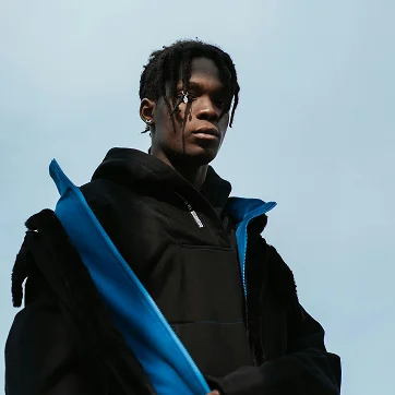 Young man wearing a black hoodie and jacket with blue accents, standing outdoors against a light blue sky.