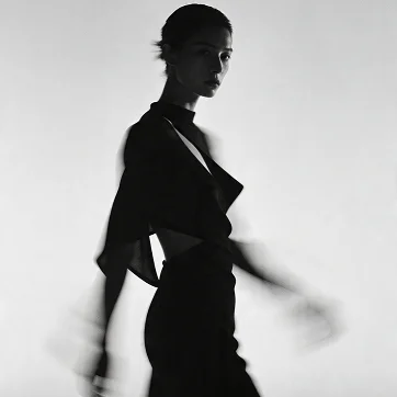 Black and white fashion portrait of a woman in a flowing black outfit, captured in motion against a light background.