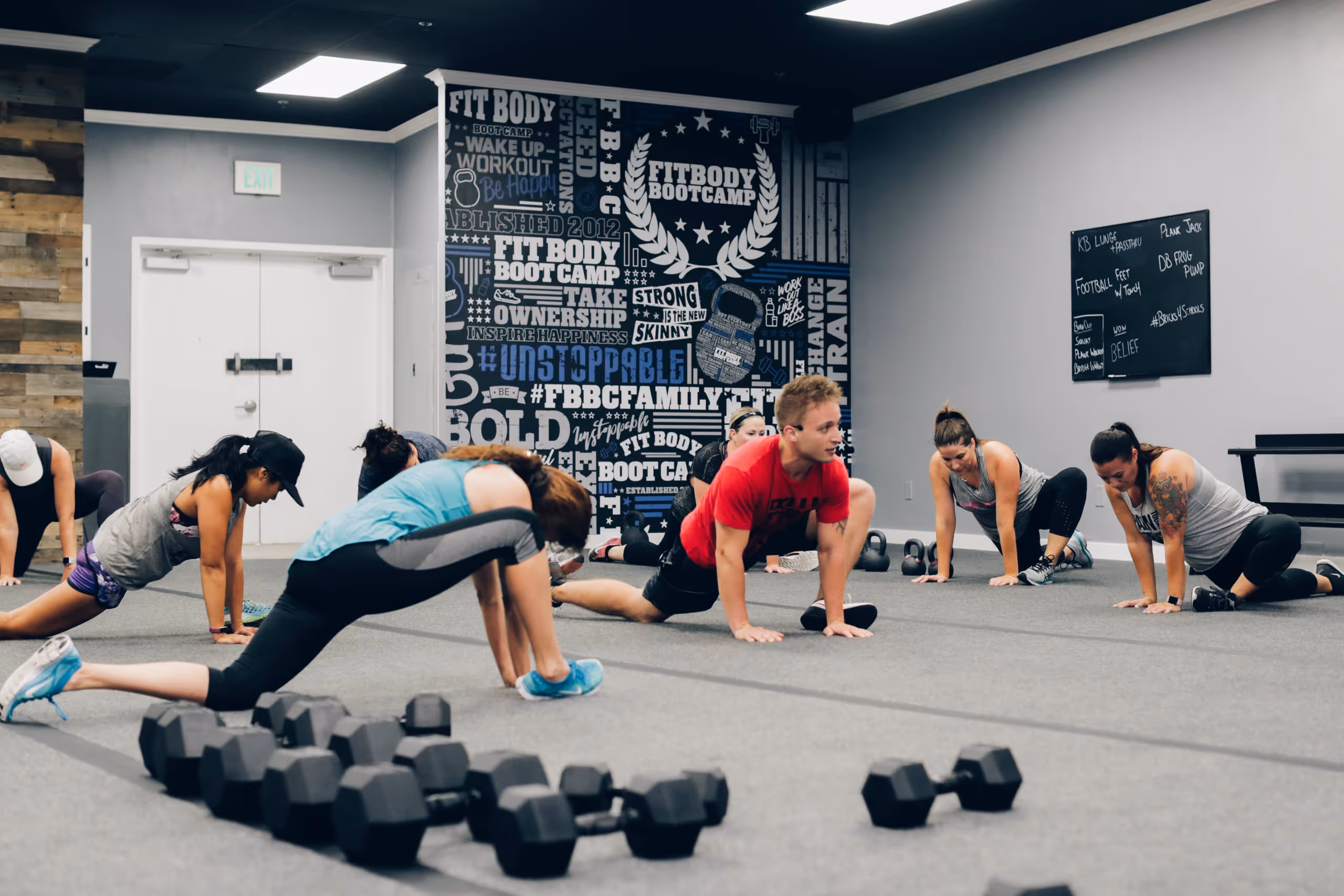 Group of people stretching on the floor in a fitness bootcamp studio with dumbbells in the foreground.