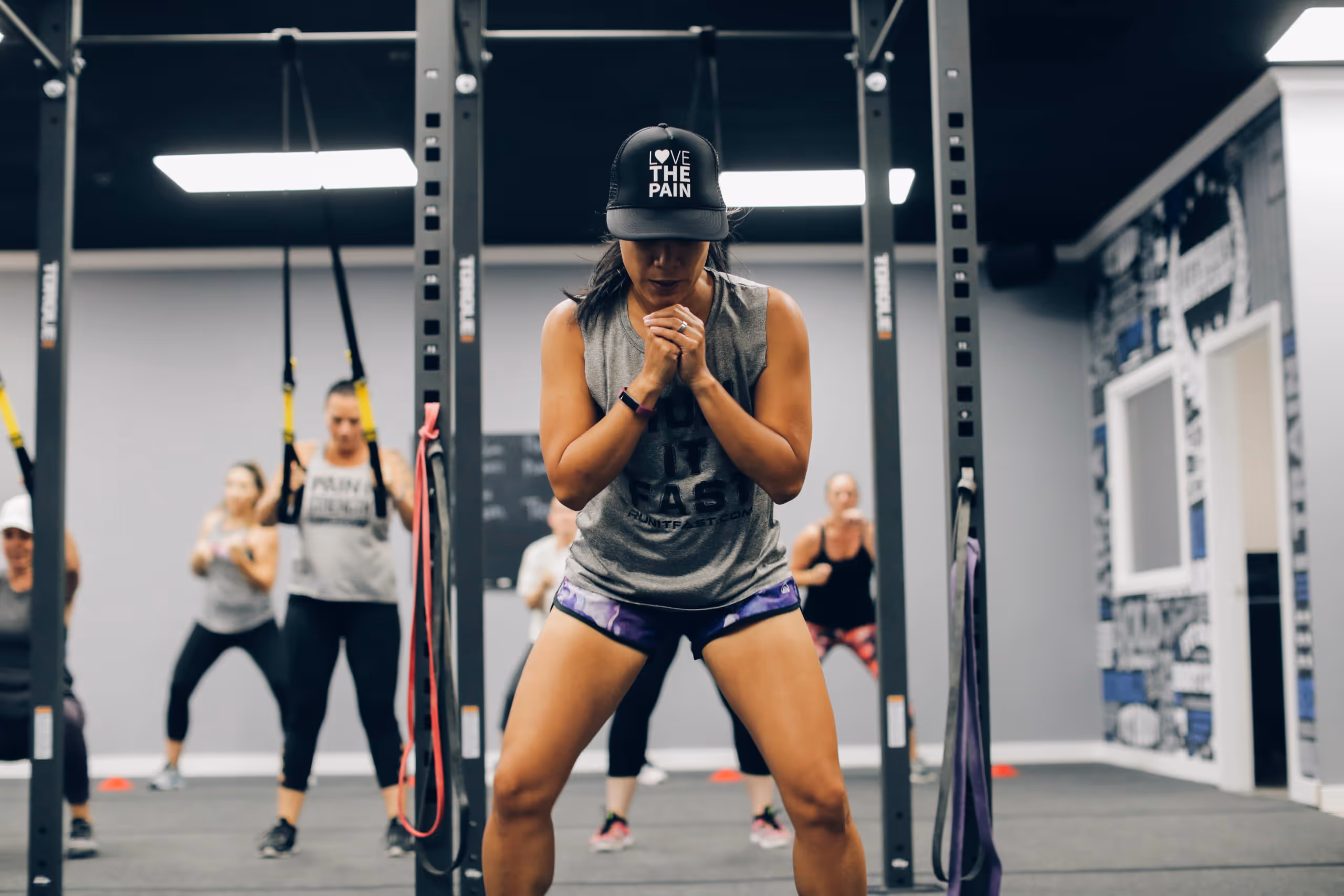 Woman wearing a cap with 'LOVE THE PAIN' text leading a group exercise session in a gym.