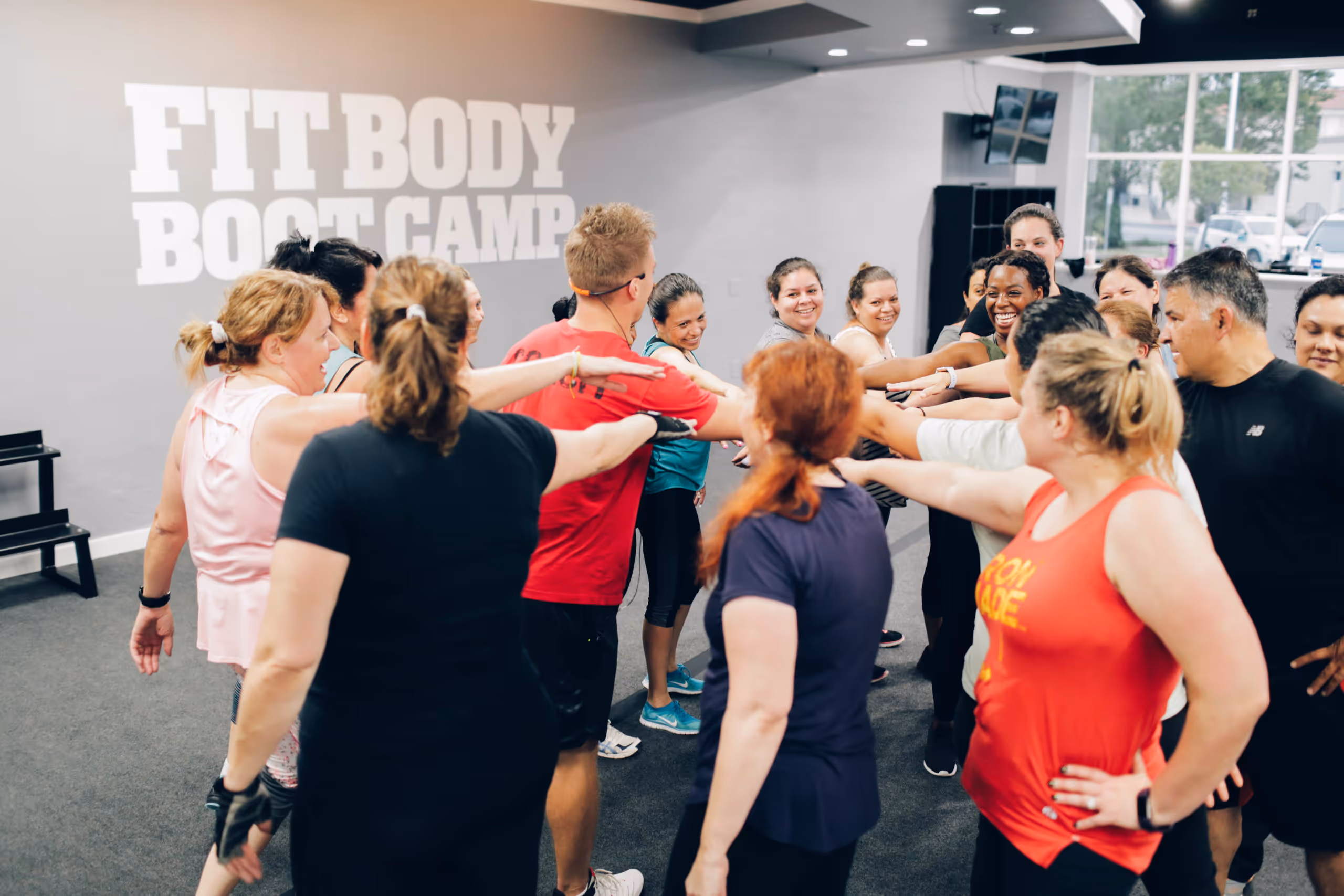 Group of diverse adults in workout clothes standing in a gym with hands together in a team huddle under a wall sign that reads 'FIT BODY BOOT CAMP'.