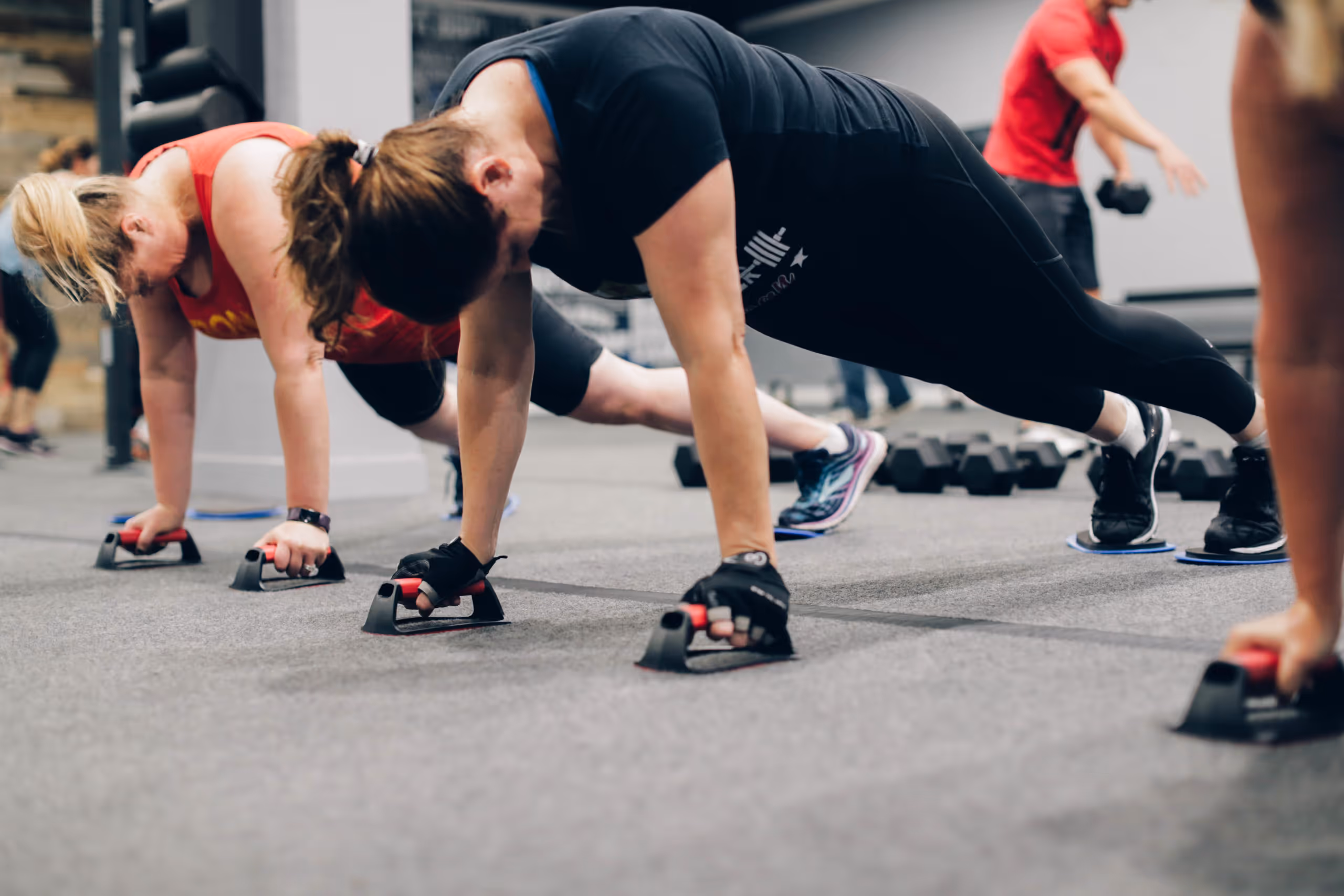 Group of people in a gym performing push-ups using push-up bars on a gray carpeted floor.