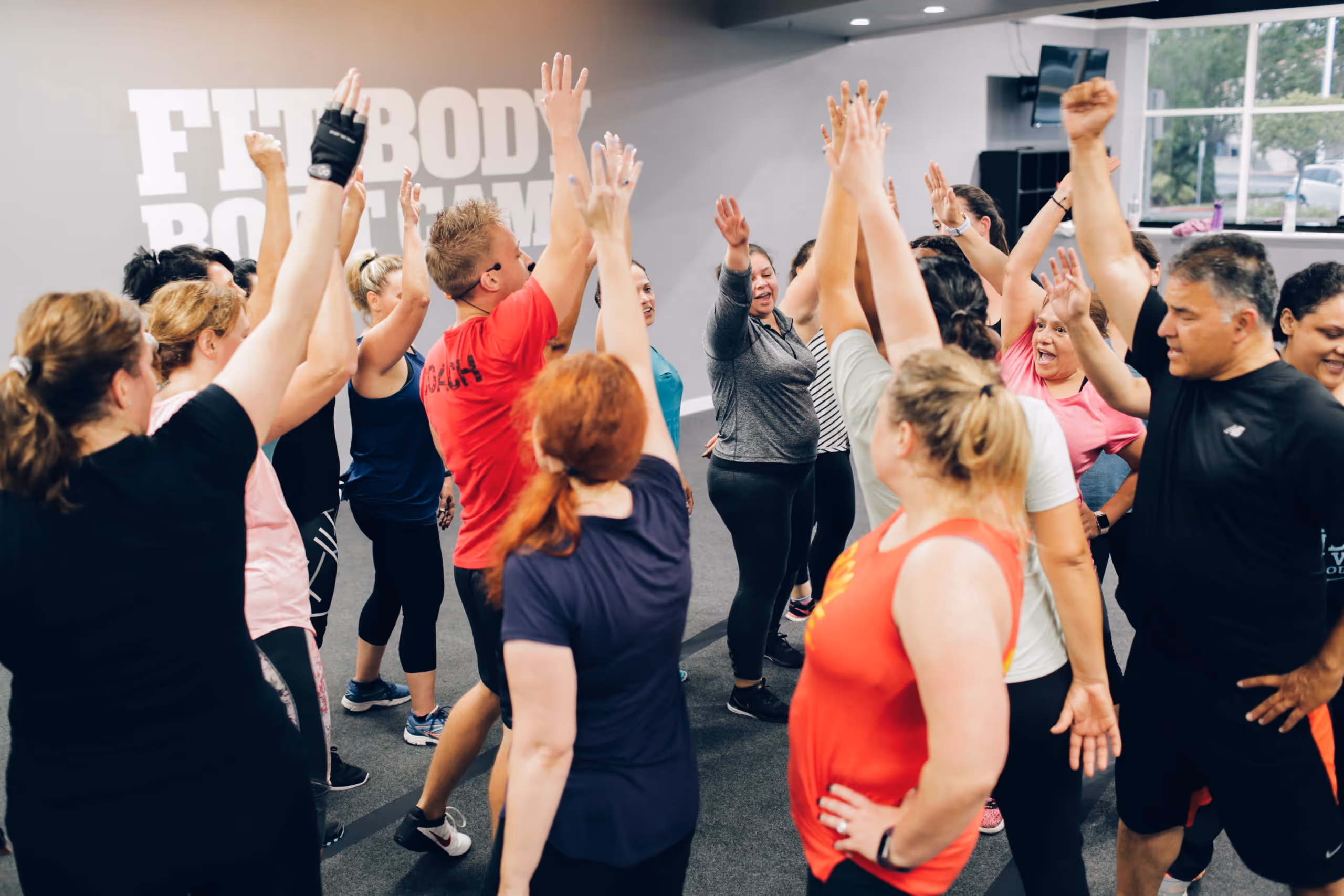 Group of diverse adults in workout clothes raising their arms together in a gym with 'Fit Body Boot Camp' on the wall.
