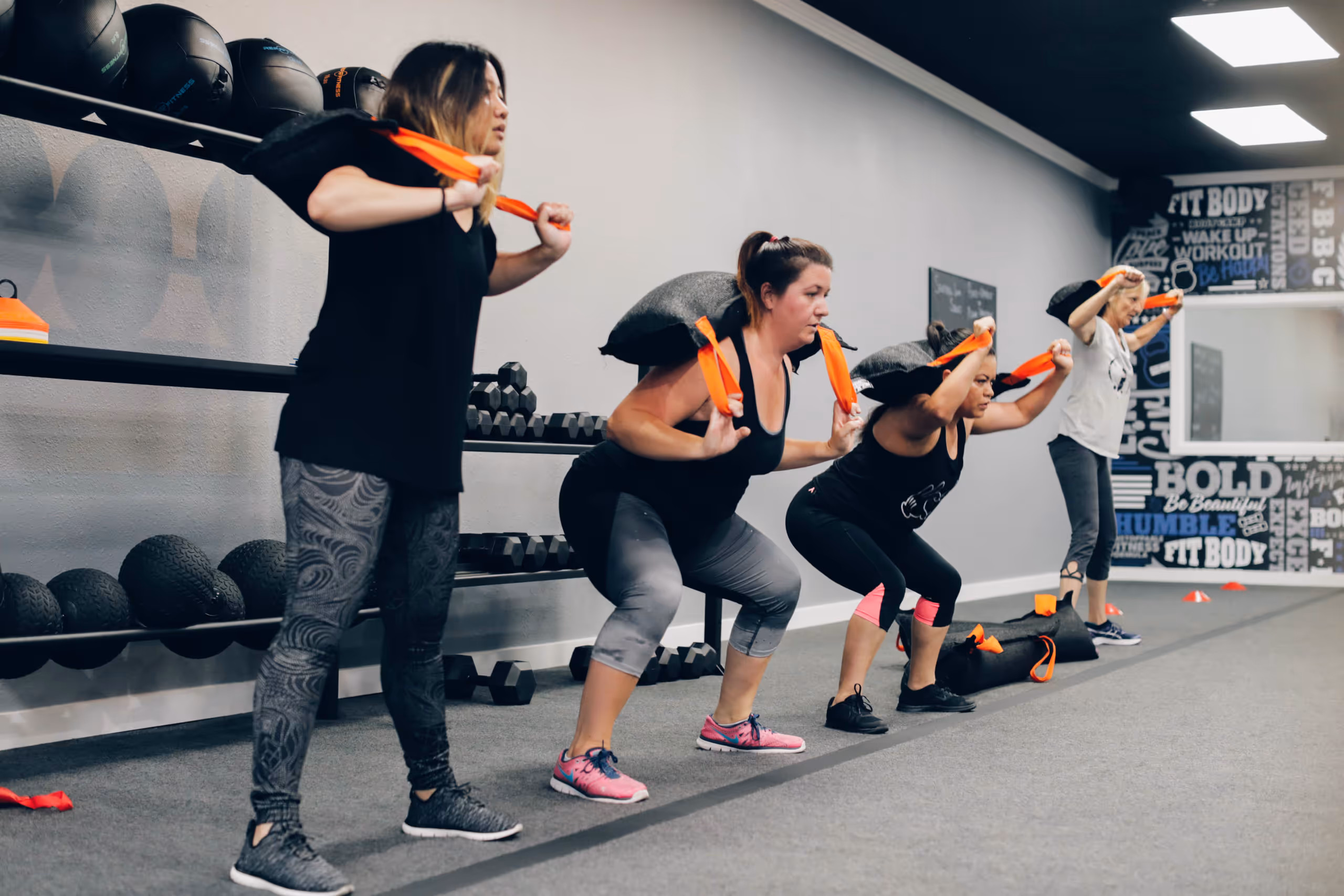 Four women exercising in a gym performing weighted squats with sandbags on their shoulders.