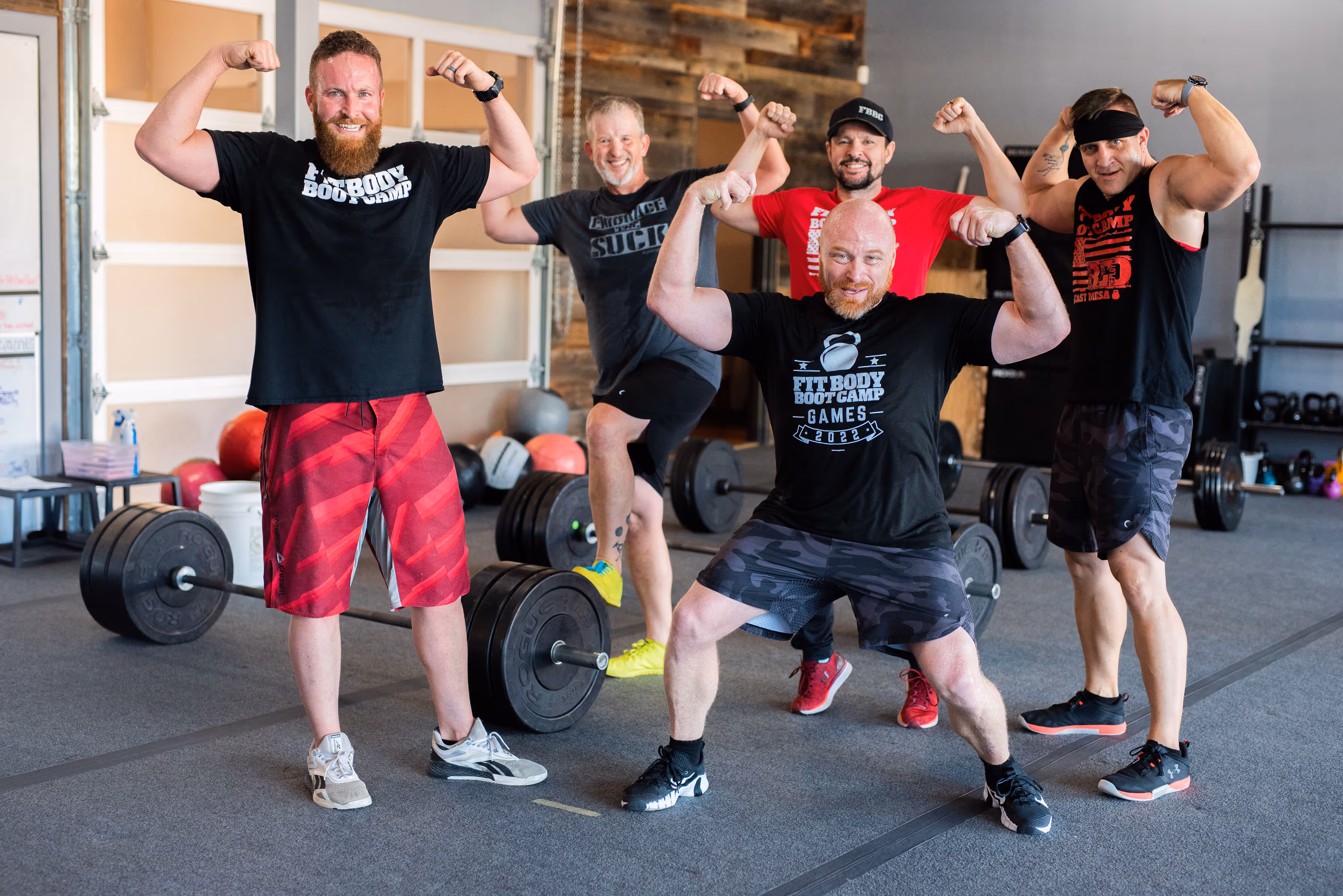 Five men in a gym flexing their arm muscles posing in front of barbells and fitness equipment.
