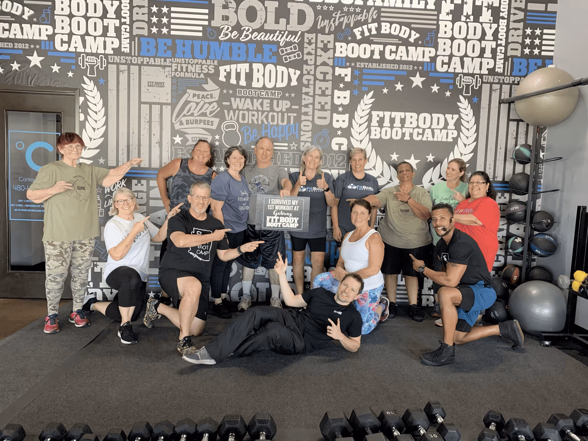 Group of people smiling and posing in a gym with a wall graphic reading Fit Body Boot Camp, one holding a sign that says 'I survived my 1st workout at Fit Body Boot Camp.'