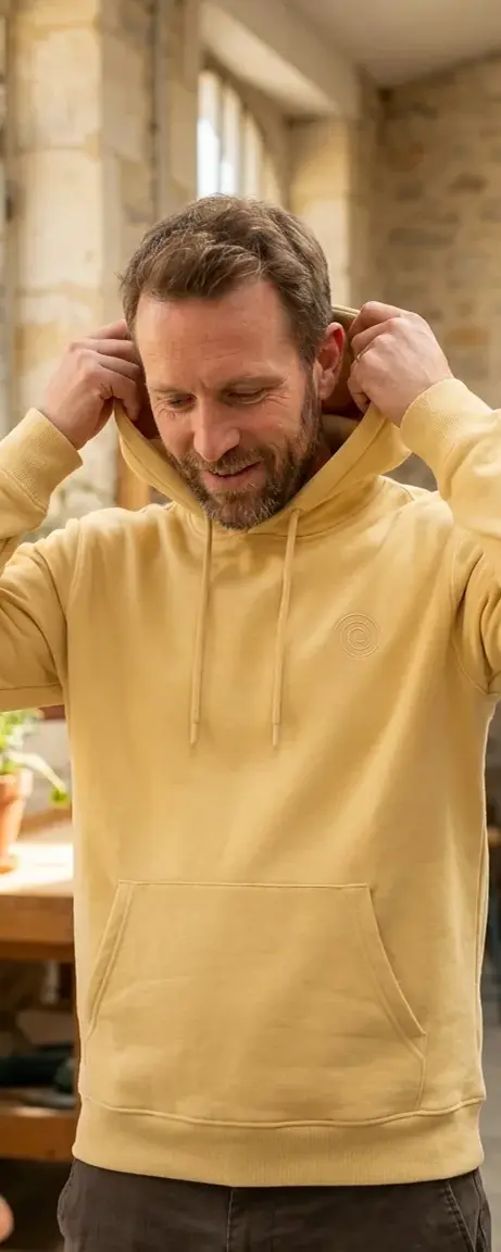 Homme ajustant la capuche d'un sweat jaune personnalisé avec logo ton sur ton, dans un intérieur en pierre — LOGOBJET