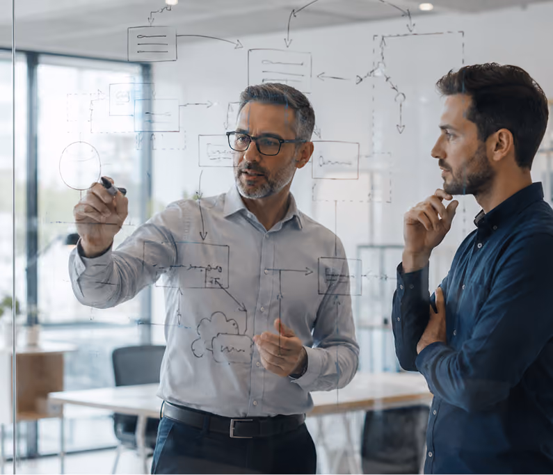 Two men in an office, one is drawing a flowchart with arrows and boxes on a transparent board.