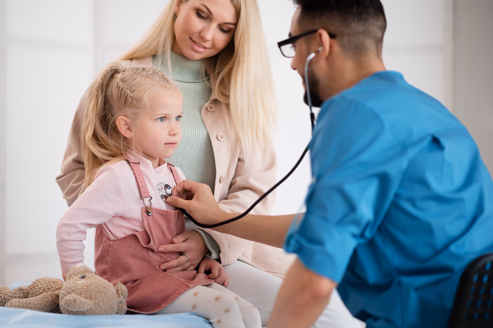 Doctor in blue scrubs listening to a young girl's heartbeat with a stethoscope as her mother holds her on an exam table - NetSuite for Healthcare