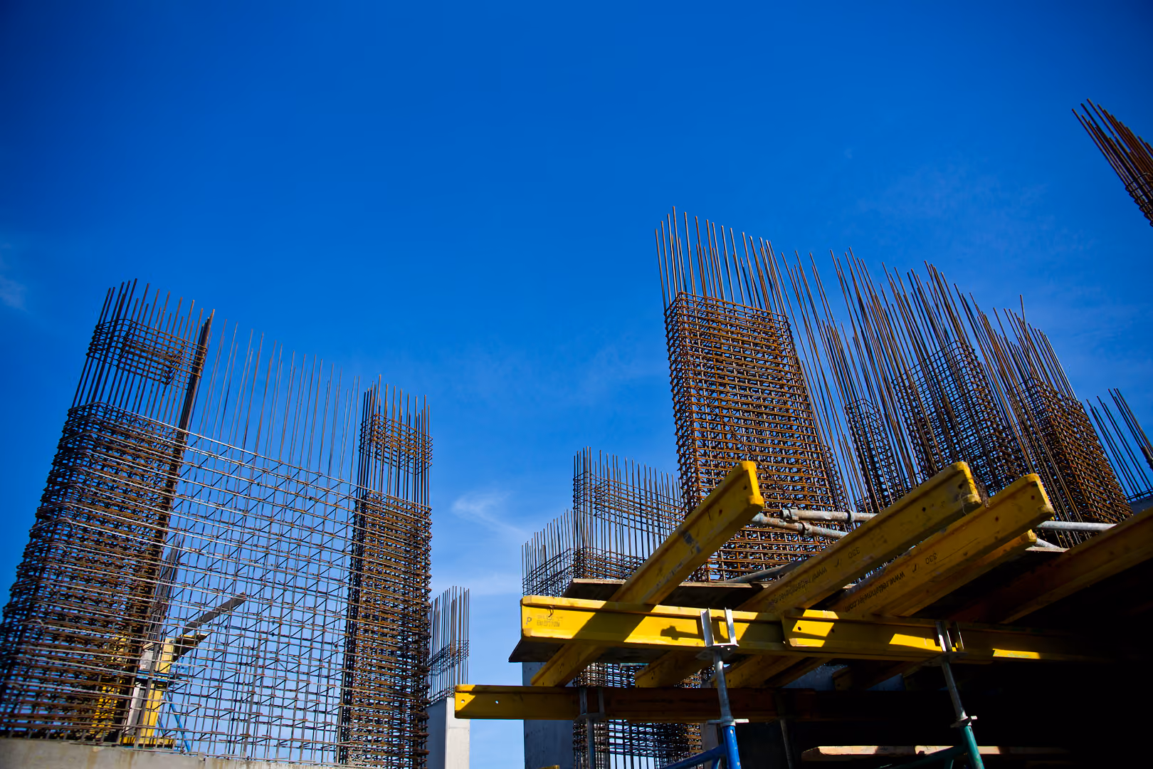 Low-angle view of steel rebar columns and yellow concrete formwork rising against a clear blue sky on a construction site - NetSuite for Construction