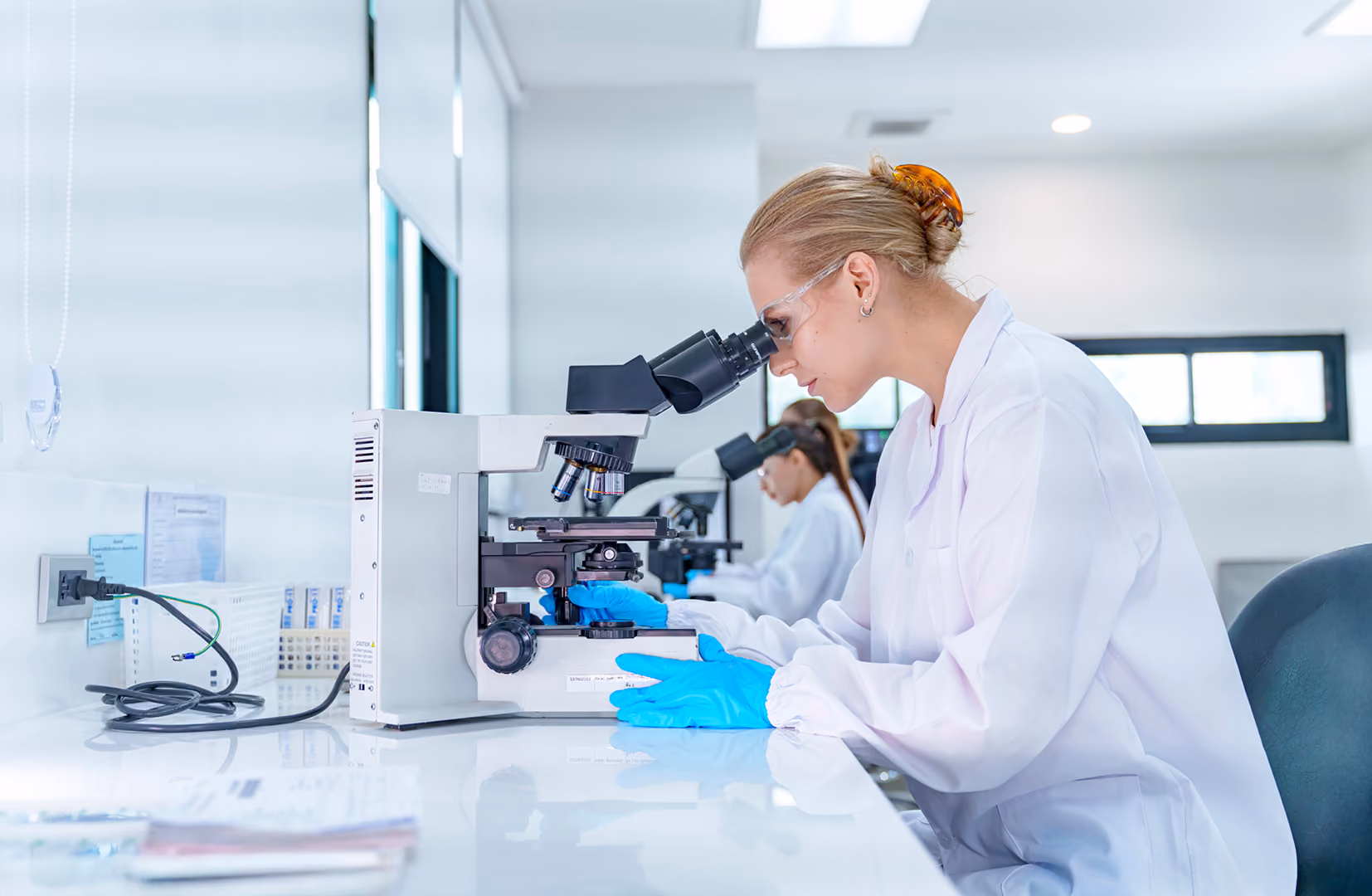 Scientists in white lab coats and blue gloves looking through microscopes at a bench in a bright laboratory - NetSuite for Life Sciences