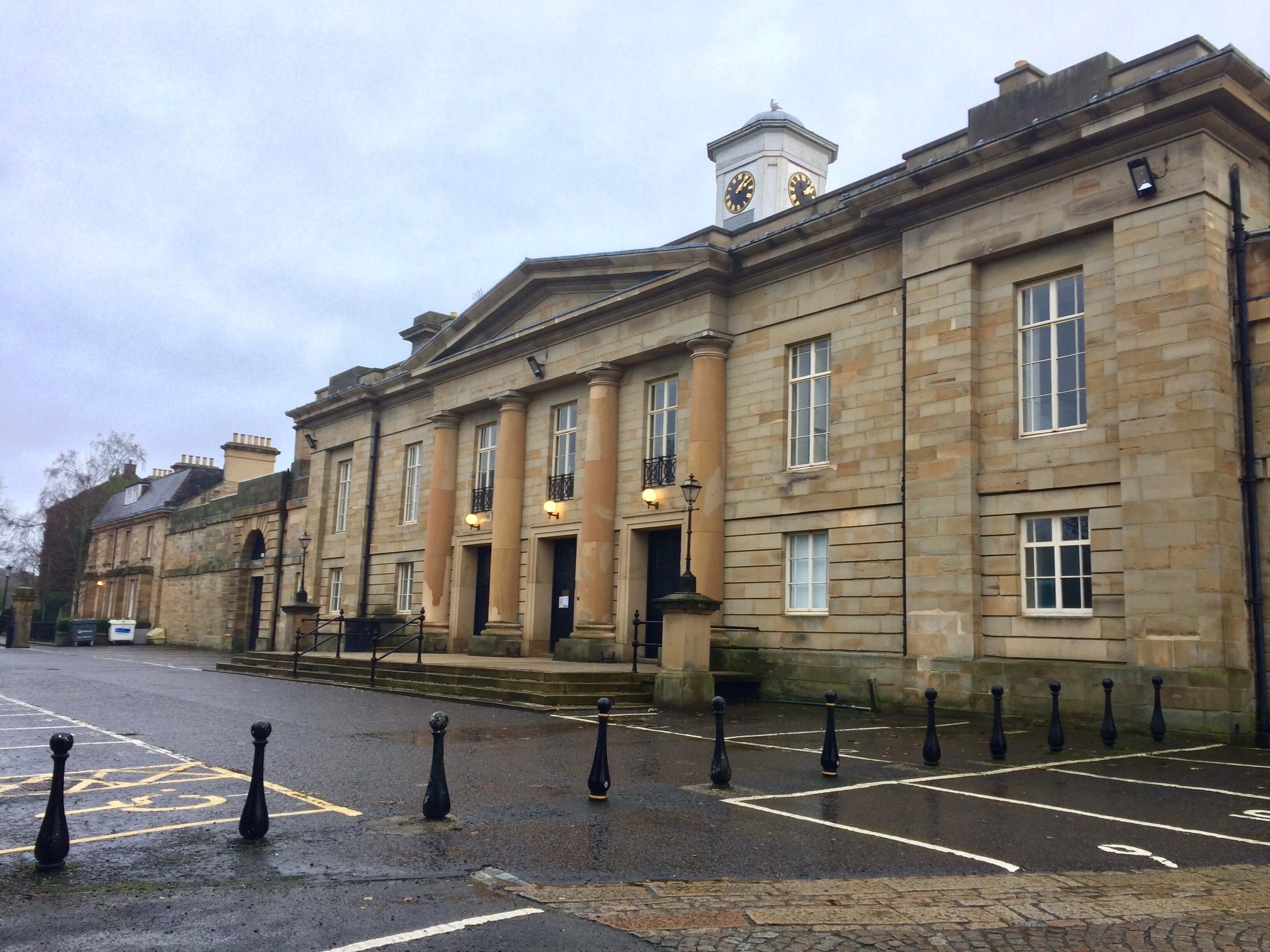 Stone building with large columns, clock tower, and lit lamps on a cloudy day.