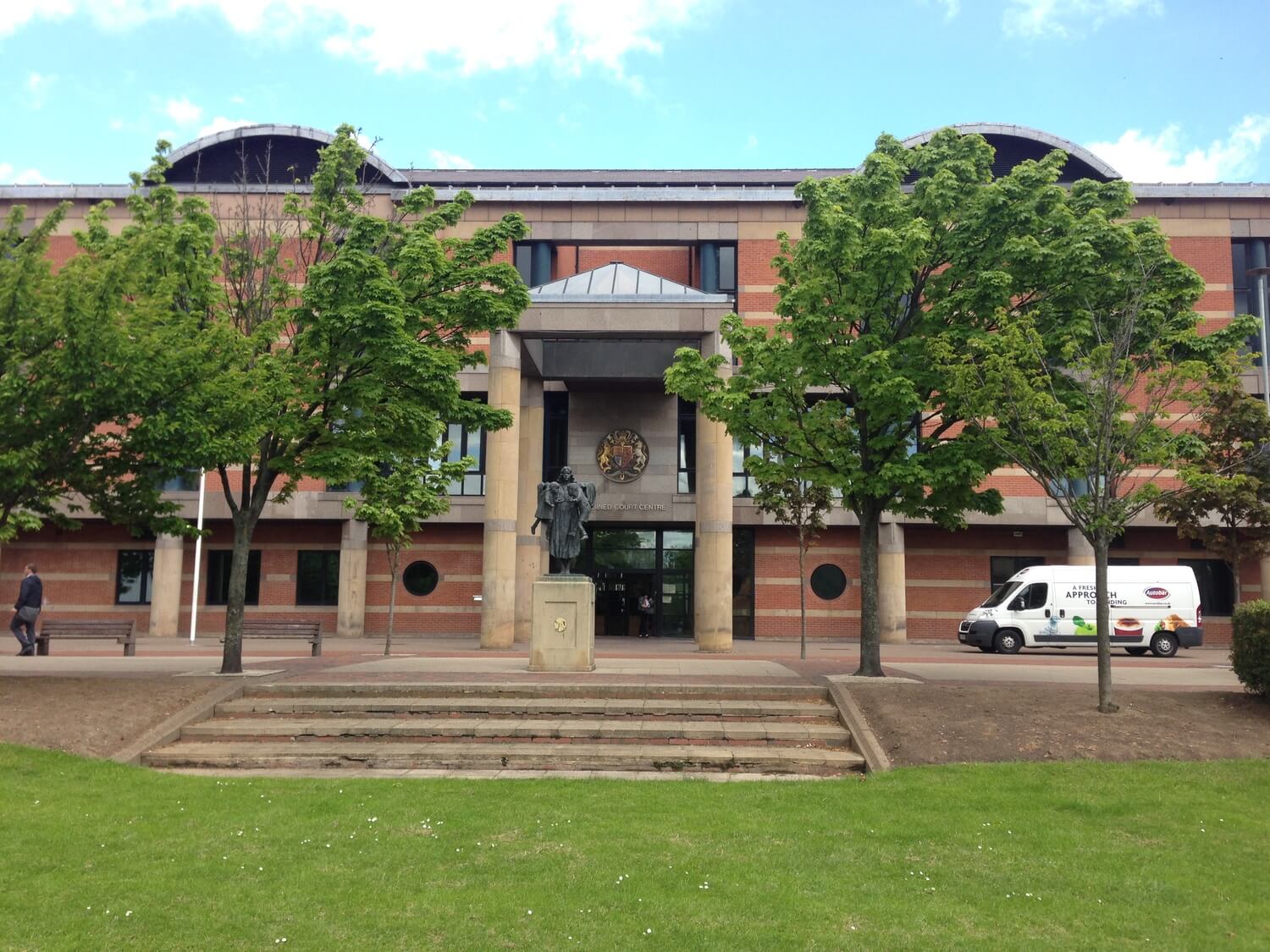 Front view of a courthouse building with trees, a statue, and a delivery van outside.