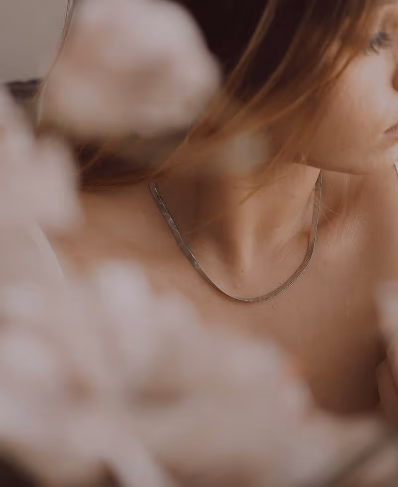Close-up of a woman wearing a thin silver necklace with blurred flowers in the foreground.