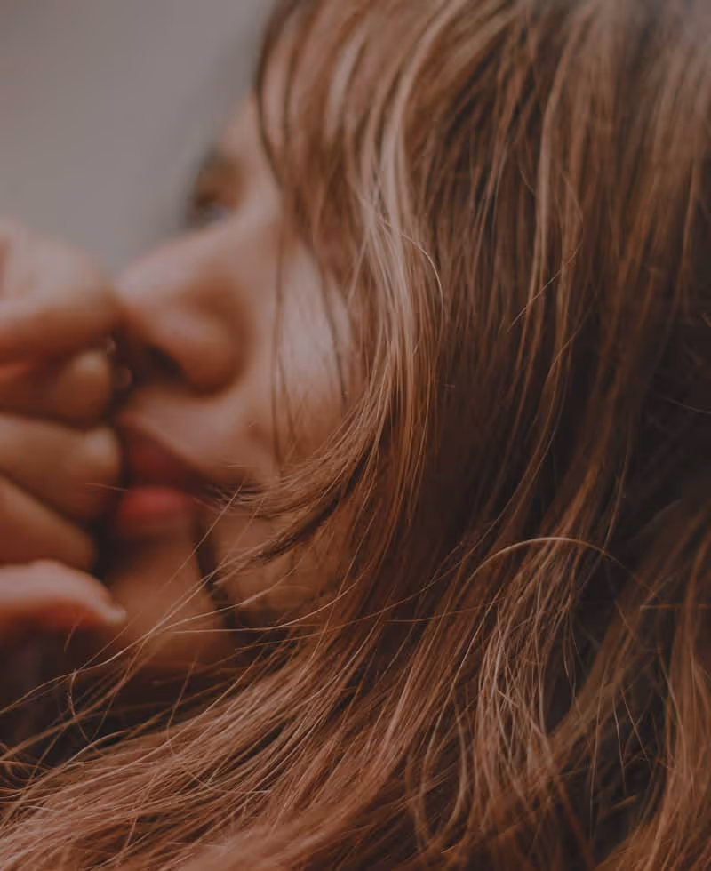 Close-up of a woman with long brown hair resting her hand near her face, looking contemplative.