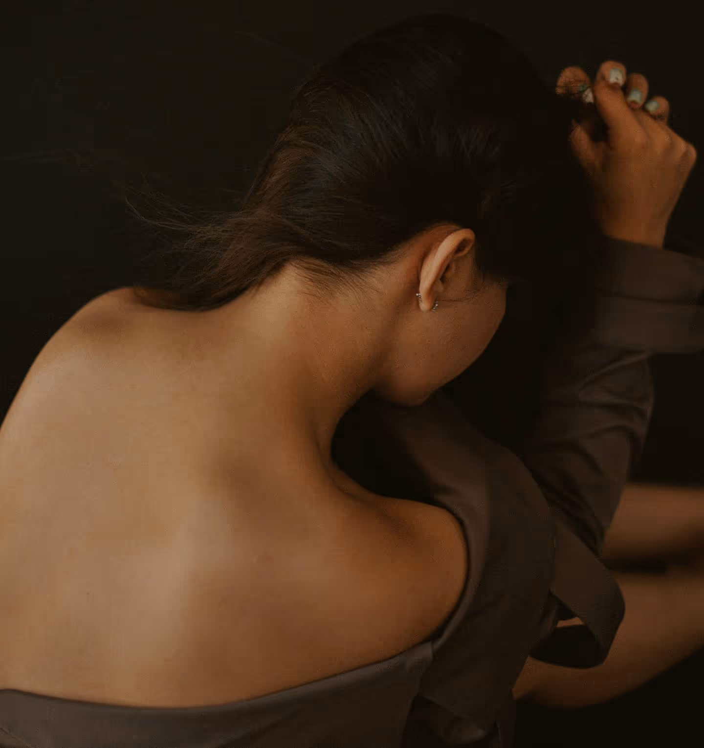 Woman with dark hair tied up, turned away showing bare back and brown off-shoulder garment.