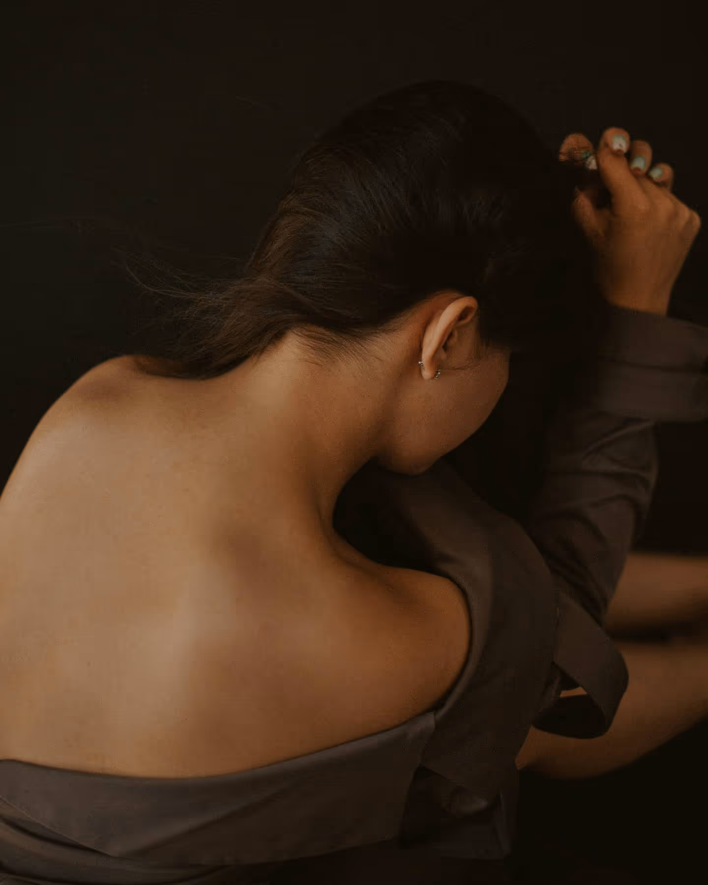 Woman with dark hair pulling it back, showing bare back and shoulder against black background.