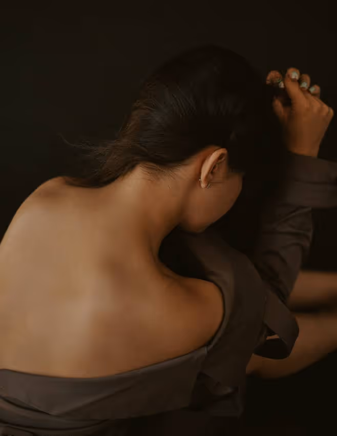 Woman with dark hair facing downward, showing bare back and nape in a dark setting.