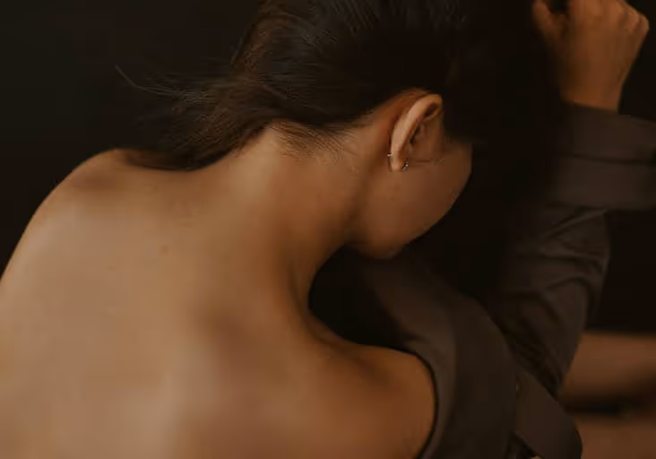 Frau mit dunklen Haaren, halb entkleidet, lehnt mit Arm an schwarze Wand.