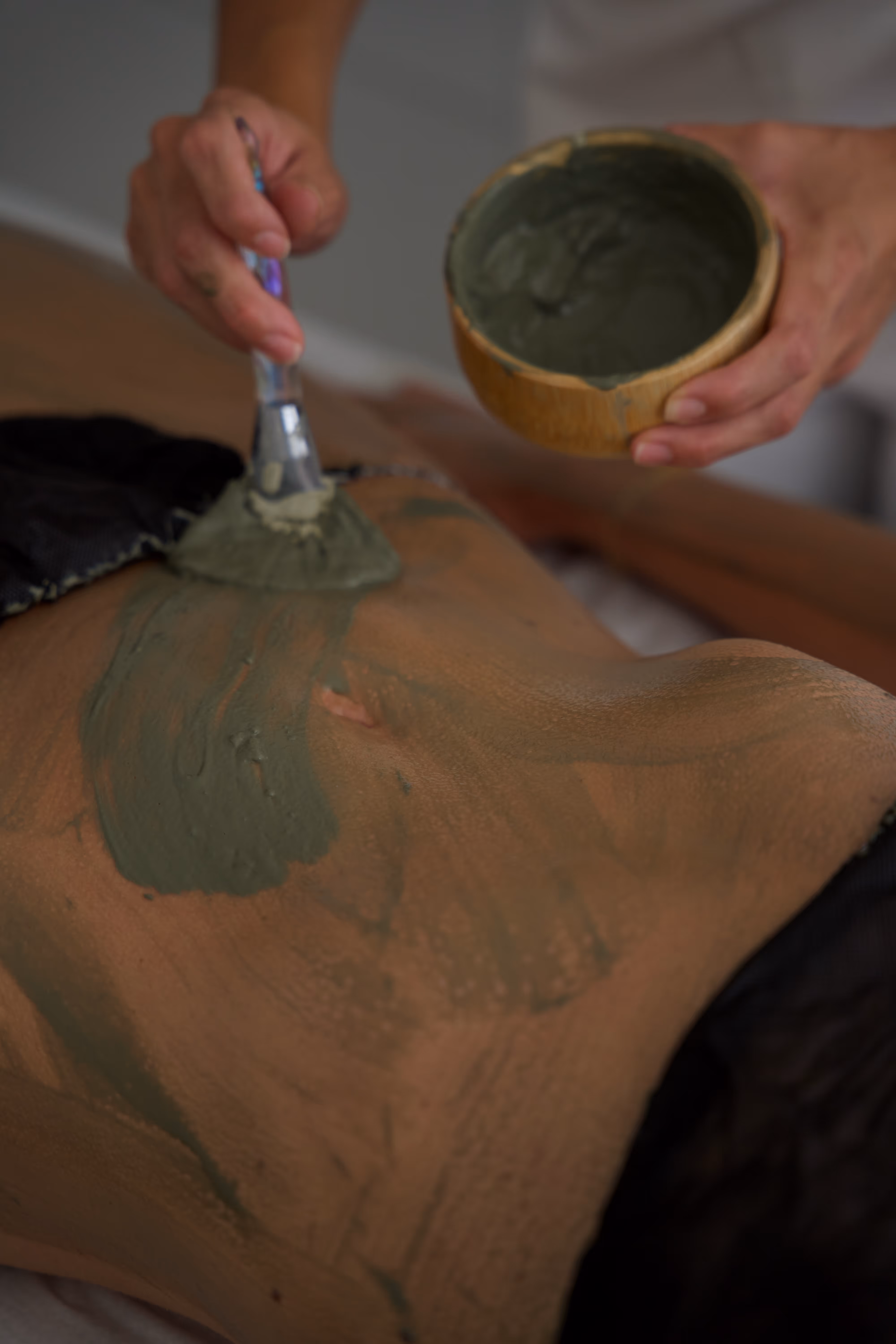 Person erhält eine Massage mit grünem Schlamm, auf Bauch aufgetragen mit Pinsel.