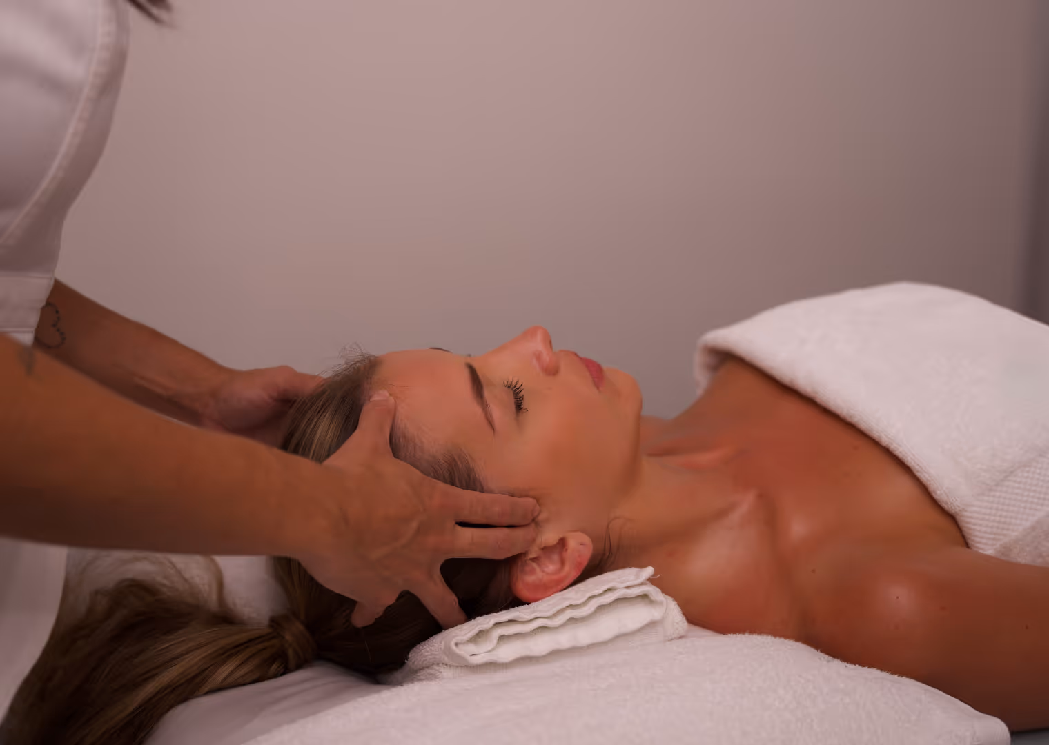 Frau erhält Kopfmassage, liegt entspannt mit geschlossenen Augen auf weißer Liege mit Handtuch