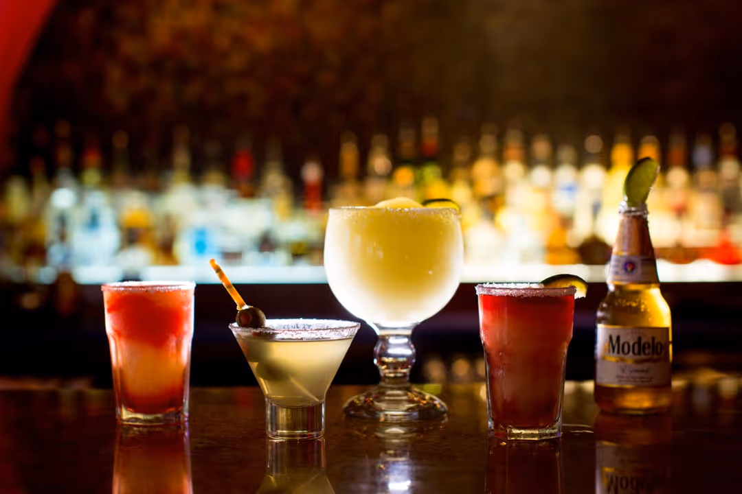 Five assorted alcoholic drinks on a bar counter with a blurred liquor shelf in the background.