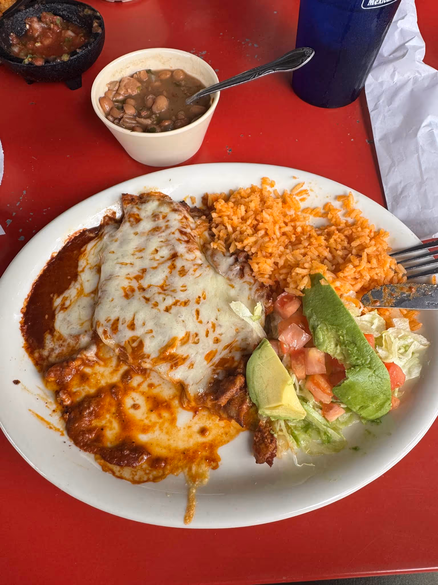 Plate with melted cheese enchiladas, Mexican rice, lettuce, tomato, avocado, and beans in a cup on red table.