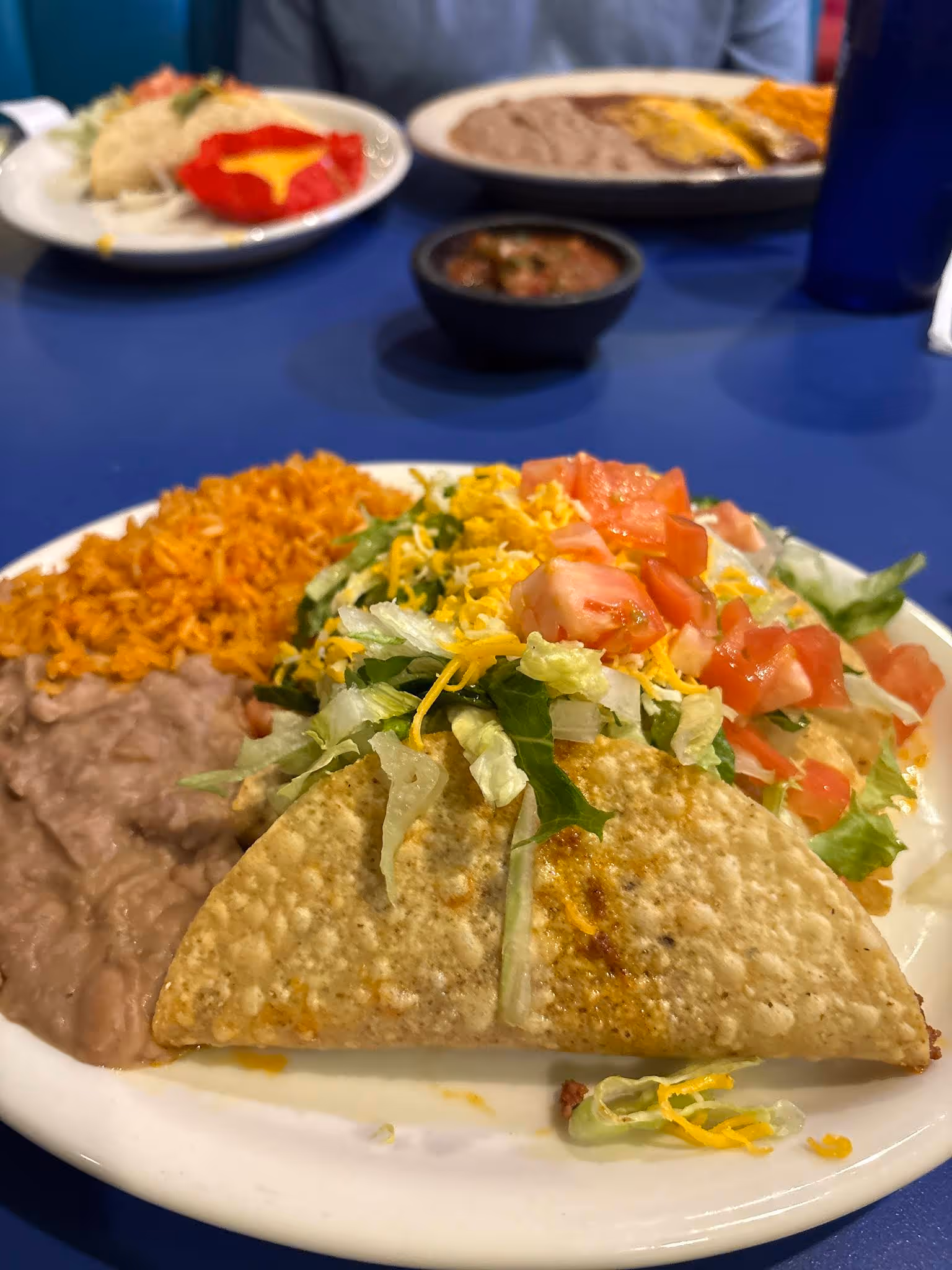 Plate with two tacos topped with lettuce, tomatoes, and cheese, served with refried beans and Spanish rice.