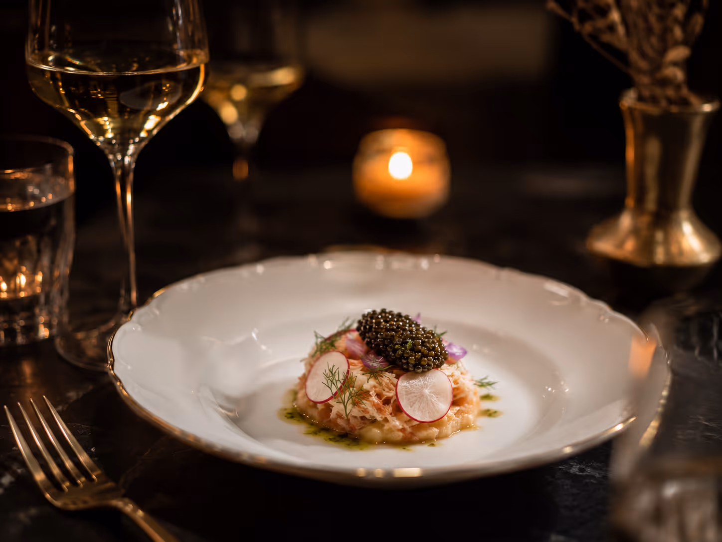 Gourmet dish with caviar, radish slices, and herbs on a white plate with wine glass nearby.