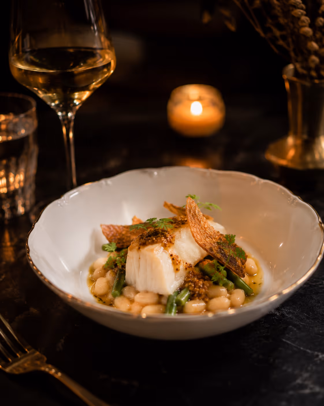 Seared fish on white beans and green vegetables with crispy garnish and mustard sauce in a white bowl.