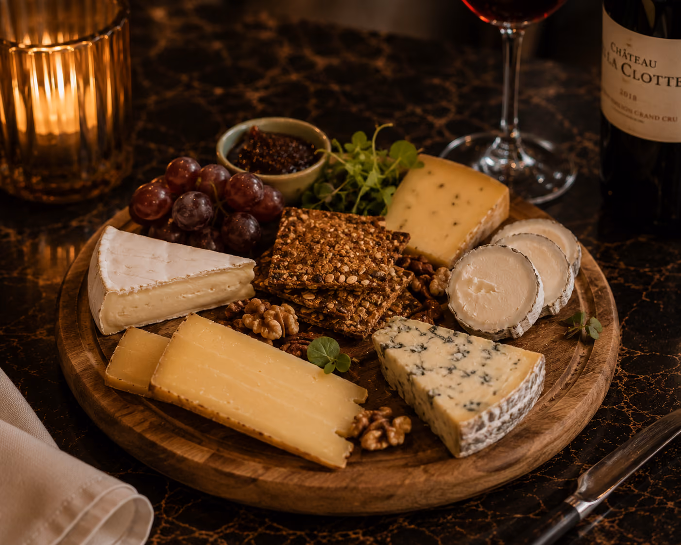 Wooden board with various cheeses, grapes, nuts, crackers, and a glass of red wine on a marble table.